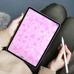 May include: A black tablet displaying a pink floral wallpaper with the time 11:04. A white stylus is held nearby. The tablet is held on a person's lap, with a beige sweater and olive green pants visible.