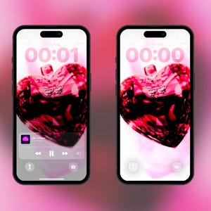 Könnte beinhalten: Zwei Smartphones nebeneinander, jedes mit einem Sperrbildschirm, der eine große, facettierte, tiefrote herzförmige Edelsteingrafik zeigt. Der linke Bildschirm zeigt eine Musik-Player-Oberfläche, beide zeigen die Uhrzeit 00:01 und 00:00.