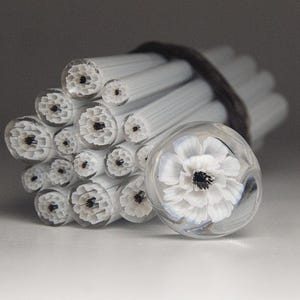 May include: A bundle of white glass rods bound with a dark band, next to a clear glass orb with a white flower and black center. The rods have a textured, floral-like pattern on their ends. The orb is round and smooth, with the flower design inside.