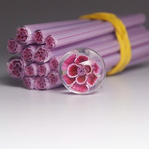 May include: A collection of lavender glass rods, secured with a yellow band, and a clear glass marble featuring a detailed pink and red flower. The ends of the rods display a floral pattern.
