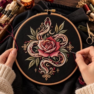 Puede incluir: Un bastidor de bordado circular de madera con tela negra que presenta un diseño detallado de una rosa roja central rodeada por una serpiente de color crema y rojo, hojas verdes, pequeñas lunas crecientes y símbolos de cruces doradas.