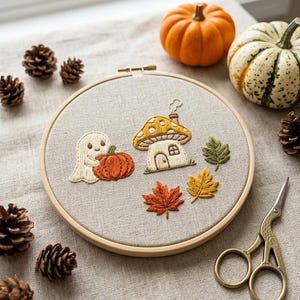 Peut inclure: Un tambour à broder contenant un tissu beige clair avec un motif brodé représentant un petit fantôme blanc tenant une citrouille orange, une maison champignon jaune avec une cheminée et trois feuilles d'automne vertes, jaunes et rouges.
