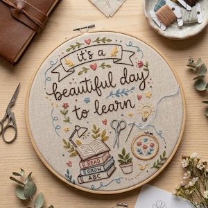 May include: An embroidery hoop with the phrase "it's a beautiful day to learn". The design includes floral and book motifs, with scissors, thread, and a notebook. A pencil and a drawing are also visible, all on a wooden surface.