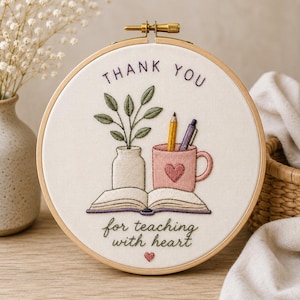 Può includere: Ricamo su telaio con le parole "THANK YOU" e "for teaching with heart". Il design presenta un vaso con piante, una tazza con un cuore, una matita e un libro aperto. Il ricamo è nei toni del verde, rosa e viola.