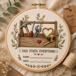 Peut inclure: Un tambour à broder représentant une boîte à outils en bois avec un marteau, un tournevis, une clé et un mètre ruban en forme de cœur. Le texte indique DAD FIXES EVERYTHING, avec des lignes pour un nom et une date sur fond crème.
