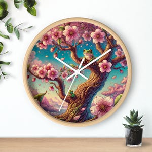 Könnte beinhalten: Eine runde Holzuhr mit weißem Zifferblatt und schwarzen Zeigern. Das Zifferblatt der Uhr zeigt eine farbenfrohe Illustration eines Baumes mit rosa Blüten und zwei grünen Fröschen. Der Baum ist vor einem blauen Himmel mit weißen Wolken platziert.