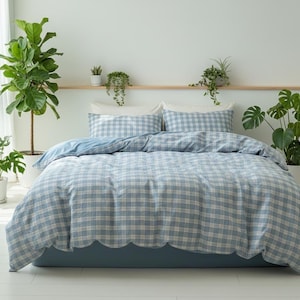 May include: A blue and white plaid duvet cover and pillow set on a bed. The bedding has a scalloped edge and is surrounded by potted plants. The room has a minimalist aesthetic with a neutral color palette.