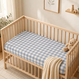 May include: A light-colored wooden crib features a fitted sheet with a blue and white gingham check pattern. A beige teddy bear sits on the mattress, and a cream-colored knitted blanket is draped over the side rail of the crib.
