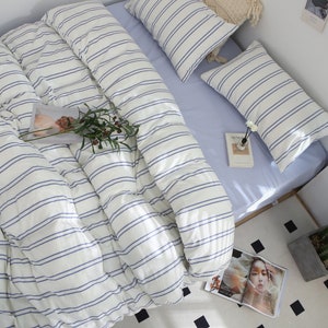 Puede incluir: Un juego de cama a rayas blancas y azules con una funda n&oacute;rdica blanca y dos fundas de almohada a juego. La ropa de cama est&aacute; dispuesta en una cama con una s&aacute;bana azul claro y una almohada a rayas blancas y azules.