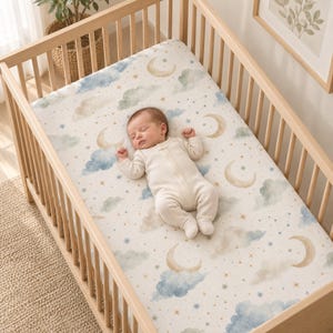 May include: A baby wearing a cream-colored long-sleeve zippered sleeper lies on a crib sheet featuring a pattern of watercolor-style crescent moons, blue and gray clouds, gold stars, and thin line constellations on a white background.