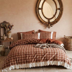 May include: A bedding set featuring a rust-colored and white plaid pattern with a decorative white lace trim along the edges. The set includes a duvet cover and matching pillow shams, complemented by a textured, neutral-toned throw blanket.