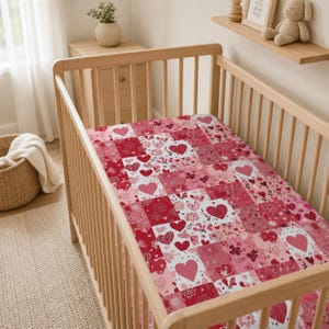 Könnte beinhalten: Ein Kinderbettlaken mit einem Patchwork-Design aus verschiedenen Rosa- und Rottönen, Herzen, Blumenmustern und feinen Linienzeichnungen. Der Stoff ist über eine Matratze in einem hellen Holzbett mit vertikalen Gitterstäben gespannt.