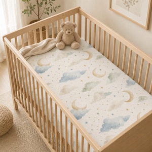 May include: A light-colored wooden crib featuring a fitted sheet with a watercolor print of crescent moons, fluffy clouds in shades of blue and beige, and small gold stars. A plush tan teddy bear sits on the mattress next to a folded beige blanket.