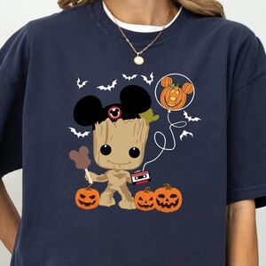 May include: Navy blue t-shirt featuring a Halloween-themed Groot character with Mickey Mouse ears and a pumpkin balloon. The design includes jack-o'-lanterns and bats. The shirt is a comfortable, oversized fit.