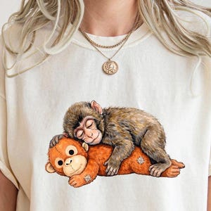 May include: Cream-colored t-shirt featuring a graphic of two cartoon monkeys. One monkey is brown and sleeping on top of an orange monkey. The design is centered on the shirt.
