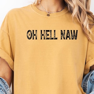 May include: A woman wearing a mustard yellow t-shirt with the text "OH HELL NAW" printed in black. She is wearing a denim jacket and denim shorts.
