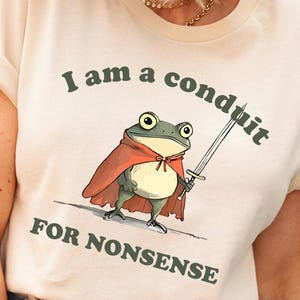May include: Cream-colored t-shirt featuring a cartoon frog in a red cape holding a sword. The text on the shirt reads "I am a conduit for nonsense" in dark green lettering. The shirt has short sleeves and a crew neck.
