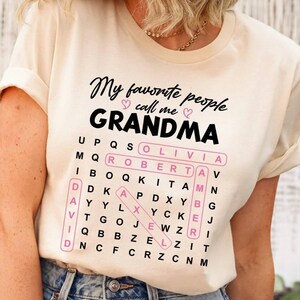 May include: Cream-colored t-shirt with the text "My favorite people call me GRANDMA" in black script. A pink-outlined word search grid includes names like Olivia, Robert, and David. The shirt is paired with blue jeans.