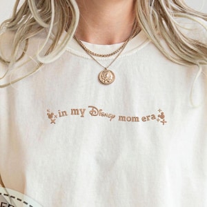 May include: A white t-shirt with the text "in my Disney mom era" in brown lettering. The shirt features a small Mickey Mouse graphic and stars.