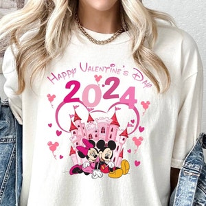 May include: White t-shirt with a pink graphic of Mickey Mouse and Minnie Mouse in front of a pink castle. The graphic also includes the text "Happy Valentine's Day 2024" with pink hearts.