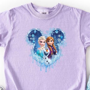 Puede incluir: Una camiseta de color lila claro con un dise&ntilde;o de Elsa, Anna y Olaf de la pel&iacute;cula Frozen, enmarcado en un dise&ntilde;o con forma de Mickey Mouse con copos de nieve. La camiseta se muestra con zapatos sin cordones a cuadros blancos y negros y pantalones cortos vaqueros.