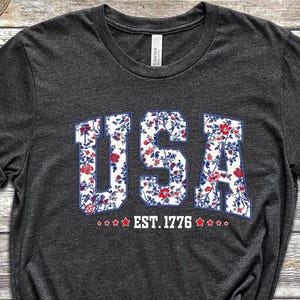 May include: Dark gray t-shirt with the word "USA" in a floral pattern, outlined in blue. Below the word "USA" is the text "EST. 1776" with star accents. The shirt is made of a soft material.