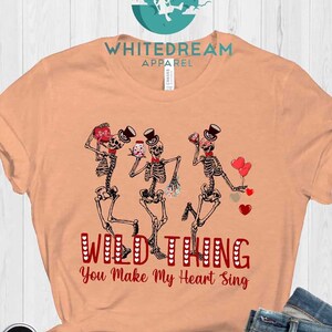 May include: A peach colored t-shirt with a graphic of three skeletons dancing and holding hearts. The text "Wild Thing You Make My Heart Sing" is printed in red below the skeletons.