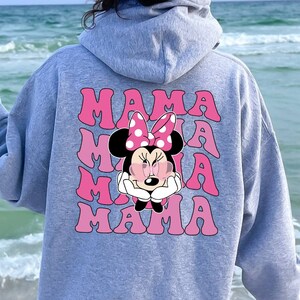以下が含まれることがあります： ピンクのグラフィックでミニーマウスがサングラスとリボンをつけているグレーのフード付きスウェットシャツ。グラフィックは4回繰り返され、ピンクの文字で「MAMA」と書いてあります。