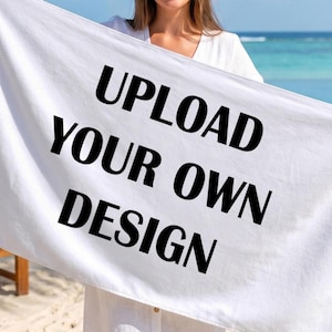 May include: A rectangular white beach towel featuring the bold black text UPLOAD YOUR OWN DESIGN centered on the fabric. The towel is made of a soft, absorbent material and is designed to be customized with personal graphics or artwork.