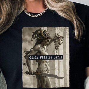 May include: Black t-shirt featuring a sepia-toned graphic of a woman holding a sword and a severed head. The text "Girls Will Be Girls" is printed across the graphic. The shirt is a crew neck style.