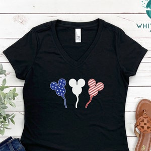 Puede incluir: Camiseta negra de cuello en V con un gr&aacute;fico de tres globos con forma de cabeza de Mickey Mouse. Los globos est&aacute;n decorados con estrellas y rayas rojas, blancas y azules, que se asemejan a la bandera espa&ntilde;ola.