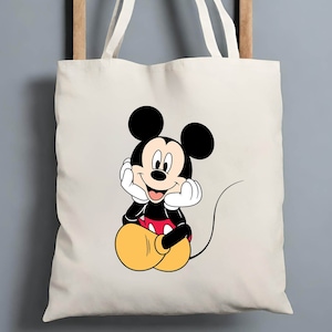 K&ouml;nnte beinhalten: Eine wei&szlig;e Canvas-Tragetasche mit einem Cartoon-Bild von Mickey Mouse, der mit den H&auml;nden am Kinn sitzt. Mickey Mouse tr&auml;gt seine charakteristischen roten Shorts und gelben Schuhe.