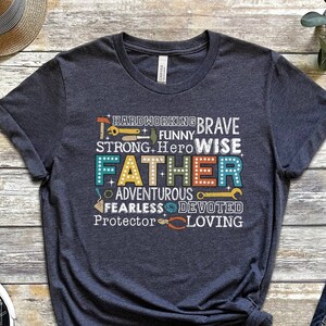 May include: Dark heather grey t-shirt with a graphic design featuring the word "Father" surrounded by words describing a father's qualities, such as "hardworking", "brave", "strong", "hero", "wise", "adventurous", "fearless", "devoted", "protector", and "loving".