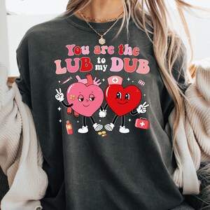 May include: A dark gray sweatshirt with a graphic of two cartoon hearts. The hearts are red with pink accents and have smiling faces. The text on the sweatshirt reads "You are the LUB to my DUB".