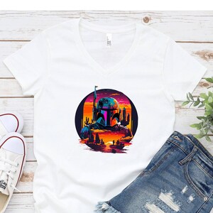 Pu&ograve; includere: Maglietta bianca a collo a V con una grafica di un personaggio di Star Wars, Boba Fett, in una scena di tramonto retr&ograve;. La grafica &egrave; in un cerchio con uno schema di colori rosa, arancione e blu.