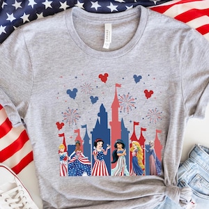 K&ouml;nnte beinhalten: Ein graues T-Shirt mit einem patriotischen Design, das eine Schlosssilhouette, Feuerwerk und Disney-Prinzessinnen in rot, wei&szlig; und blauen Kleidern zeigt. Das Shirt wird mit Jeansshorts und wei&szlig;en Turnschuhen kombiniert, mit einer amerikanischen Flagge im Hintergrund.