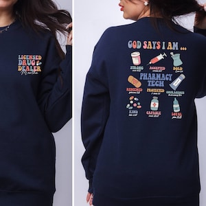 May include: A navy blue sweatshirt with a graphic design on the back that says "God Says I Am..." with a list of words and bible verses related to pharmacy. The front of the sweatshirt has the text "Licensed Drug Dealer" with the name "Martha" underneath.