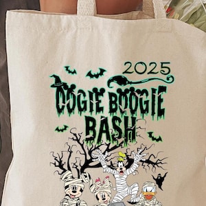 May include: A beige tote bag with a Halloween-themed graphic. The design features the text "DOGIE BOOGIE BASH 2025" in green, along with cartoon characters dressed as mummies and other Halloween elements like bats and pumpkins.