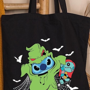 Pode incluir: Bolsa tote preta com um design de Halloween. Apresenta um personagem azul com uma fantasia verde, segurando uma boneca, com morcegos brancos e teias de aranha. A bolsa &eacute; feita de tecido dur&aacute;vel.