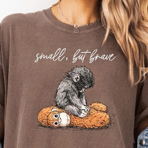 May include: A brown t-shirt featuring a graphic of a small, gray monkey sitting on a plush orange toy. The text "small, but brave" is written above the graphic in a cursive font. The shirt has a relaxed fit.