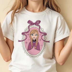 May include: Cream-colored t-shirt featuring a cartoon illustration of a character with blonde braids, wearing a purple cape, inside a pink oval frame. The frame is adorned with a purple ribbon and a beaded border. The shirt is short-sleeved.