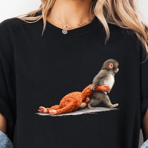 May include: Black t-shirt featuring a whimsical illustration of two monkeys. One monkey is carrying another, with the carried monkey's arms wrapped around the carrier's waist. The image is a playful design.
