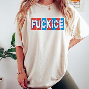 以下が含まれることがあります： 赤、白、青のブロックデザインで「FUCKICE」という言葉が入ったクリーム色のTシャツ。Tシャツは半袖でゆったりとしたフィット感です。テキストは胸の中央に配置されています。