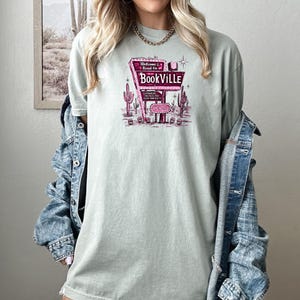 May include: A light green t-shirt with a pink graphic of a vintage neon sign that reads "Welcome & Read On Bookville". The sign is surrounded by cacti and a star.