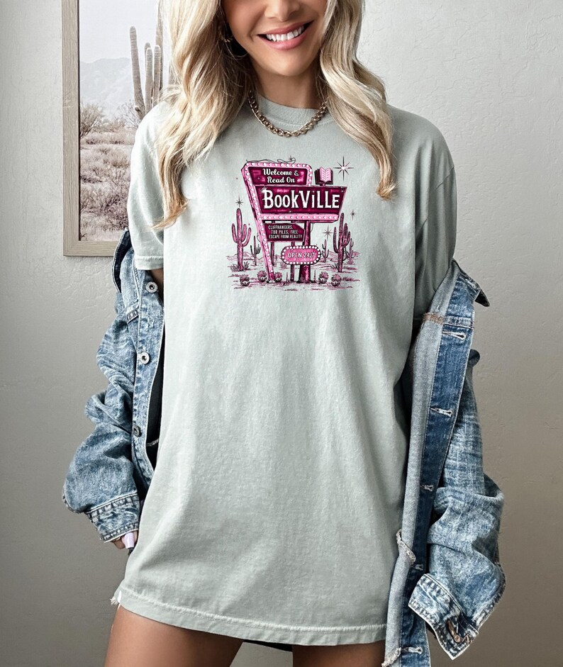 May include: A light green t-shirt with a pink graphic of a vintage neon sign that reads "Welcome & Read On Bookville". The sign is surrounded by cacti and a star.