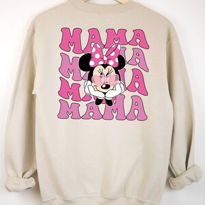 以下が含まれることがあります： ピンクと白のミニーマウスのグラフィックが描かれたベージュのトレーナー。"MAMA"という文字が4回繰り返されています。
