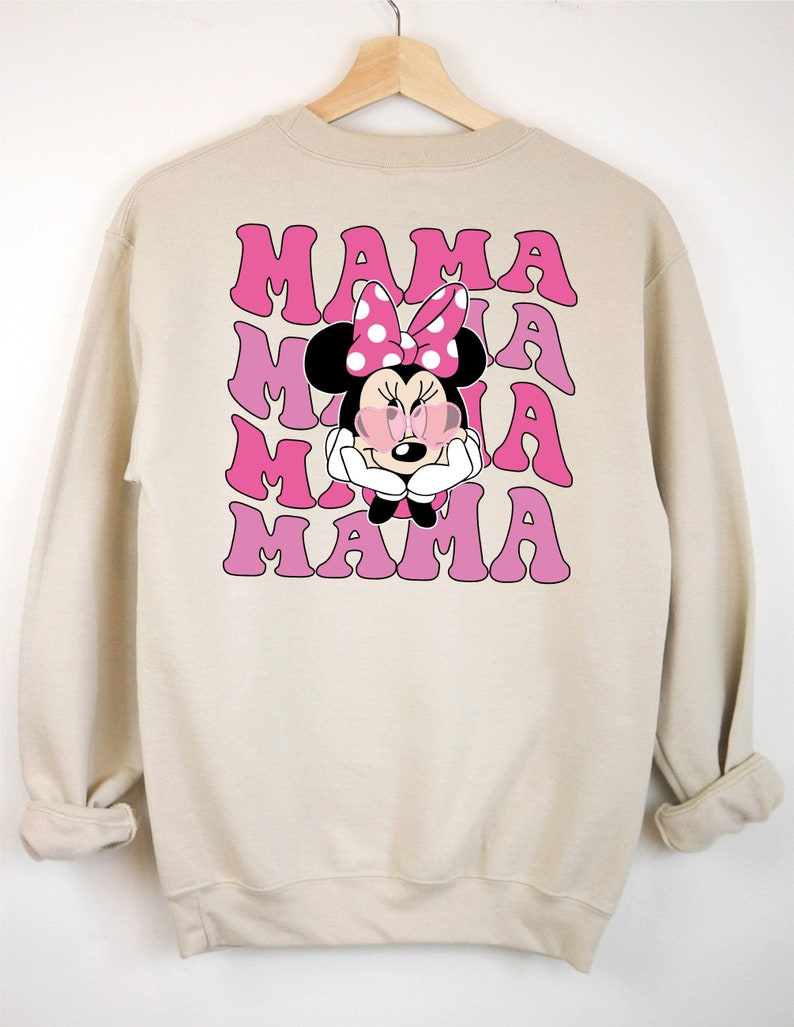 以下が含まれることがあります： ピンクと白のミニーマウスのグラフィックが描かれたベージュのトレーナー。"MAMA"という文字が4回繰り返されています。