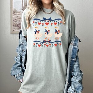 May include: A light green t-shirt featuring three cartoon ducks with blue bows. The ducks are centered on the shirt, above and below a blue and red ribbon design with hearts and flowers. The shirt is worn with a denim jacket.