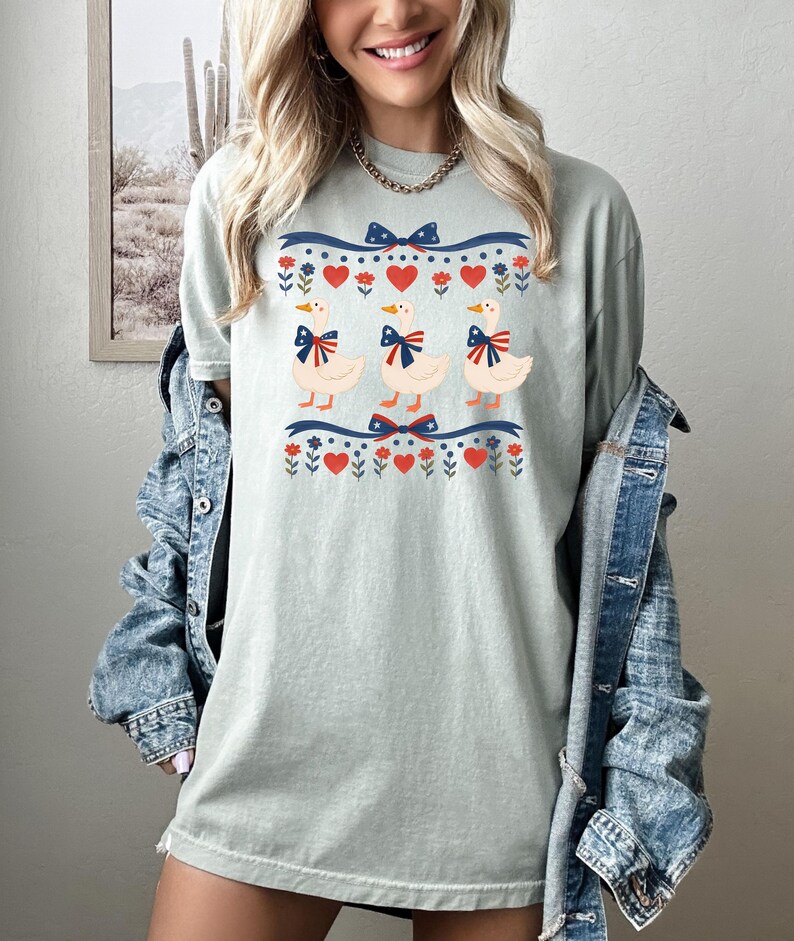 May include: A light green t-shirt featuring three cartoon ducks with blue bows. The ducks are centered on the shirt, above and below a blue and red ribbon design with hearts and flowers. The shirt is worn with a denim jacket.