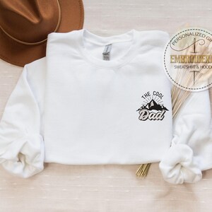 Op de afbeelding: Witte sweatshirt met de tekst "The Cool Dad" in het zwart, met een bergafbeelding. Op de achtergrond is een bruine hoed te zien. De sweatshirt heeft lange mouwen en een ronde hals. Een ronde sticker luidt "Gepersonaliseerd cadeau, Geborduurd sweatshirt & hoodie."